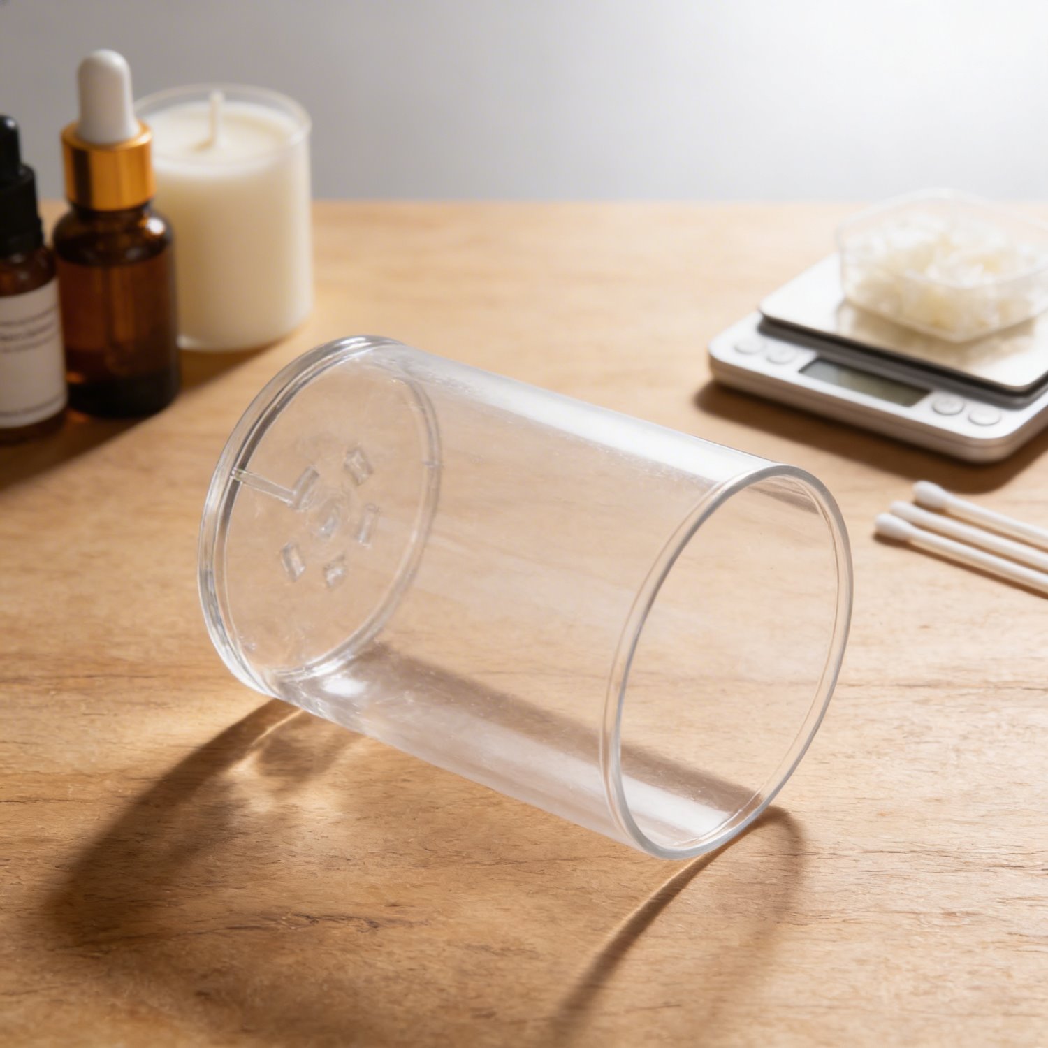 Transparenter zylindrischer Windschutzbecher aus Kunststoff, liegend auf Holzoberfläche, im Hintergrund Kerzengießzubehör wie Digitalwaage mit Sojawachs, Dochtstäbe, braune Pipettenflasche und weiße Kerze.