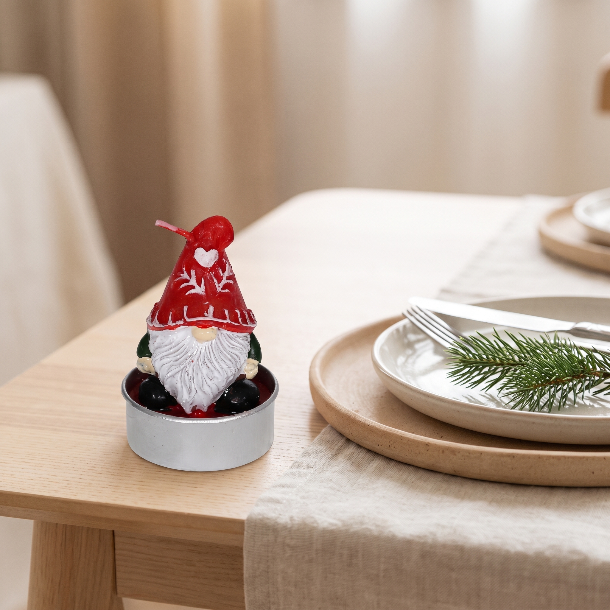 Teelicht in Weihnachtswichtel-Form mit roter Zipfelmütze und weißem Bart im silbernen Teelichthalter auf einem Holztisch, daneben weihnachtlich gedeckter Tisch mit Teller, Besteck und Tannenzweig.