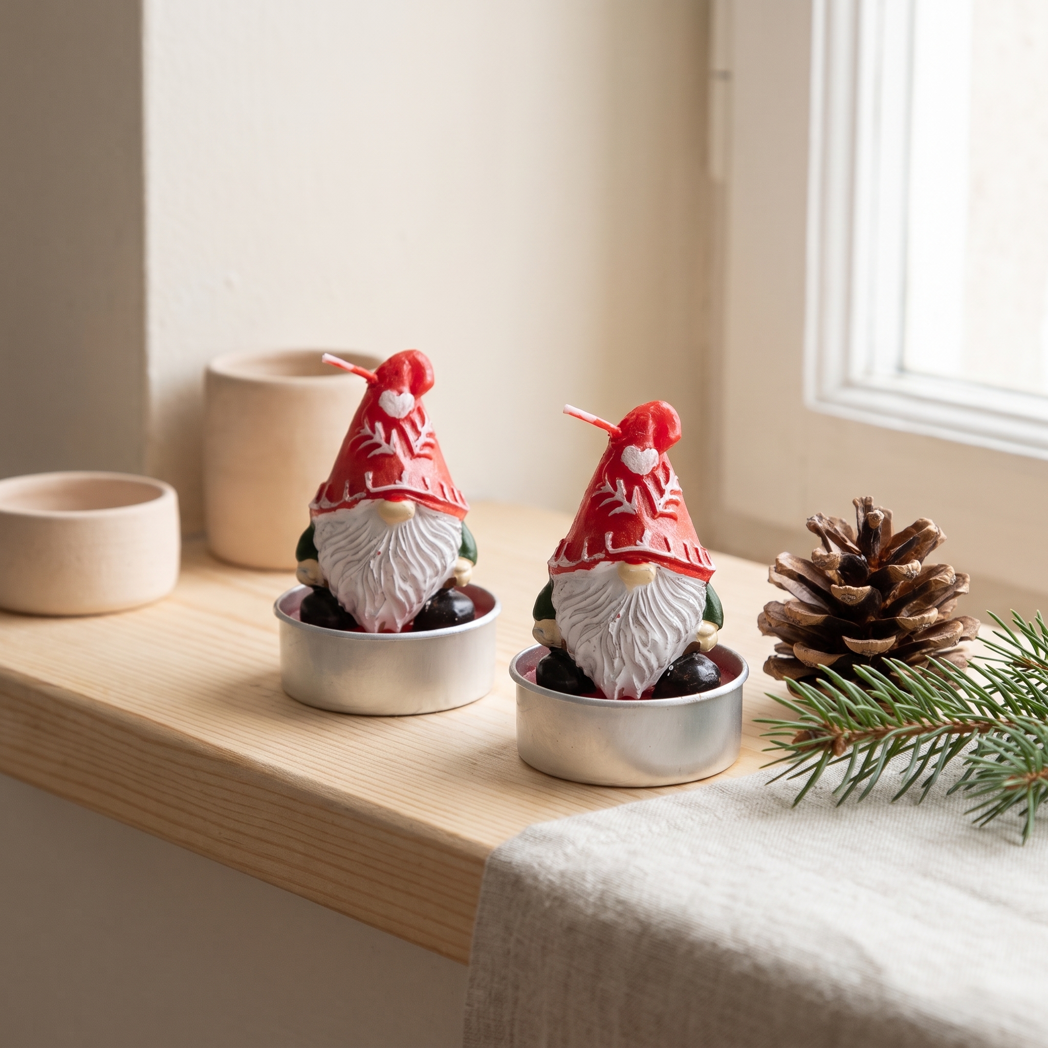 Zwei Teelicht-Kerzen in Form von Weihnachtswichteln mit roter Mütze und weißem Bart in silbernen Teelichthaltern auf einem Holzbrett, dekoriert mit Tannenzweig und Tannenzapfen