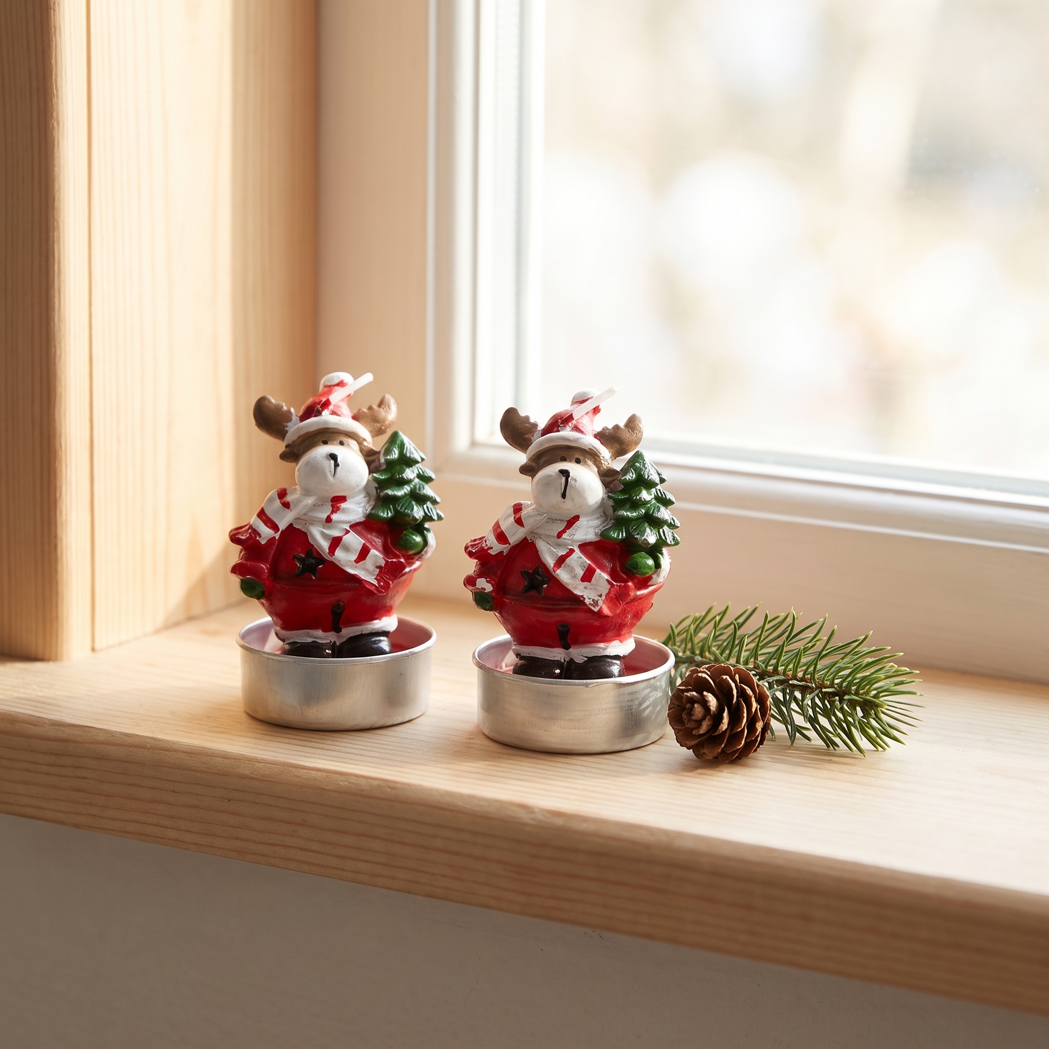 Zwei Teelichter mit weihnachtlichen Rentier-Figuren in roten Mänteln und weißen Schals, einen Weihnachtsbaum haltend, in silbernen Teelichthaltern auf einer Holzfensterbank, daneben ein Tannenzweig mit Zapfen.