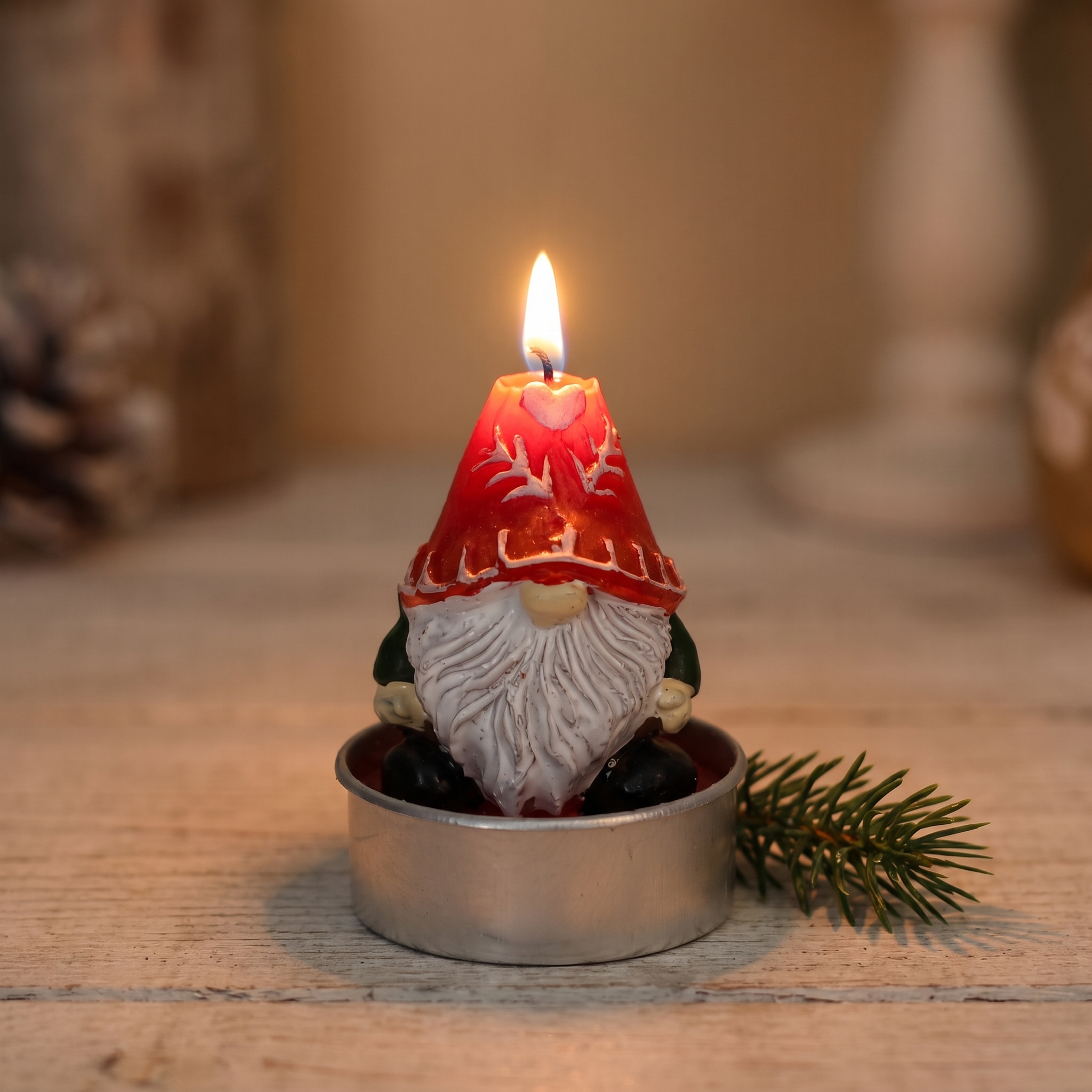 Brennendes Teelicht in Form eines Weihnachtswichtel mit roter Zipfelmütze mit Rentiergeweih-Muster und weißem Bart, in silbernem Teelichthalter auf Holzuntergrund, daneben ein Tannenzweig