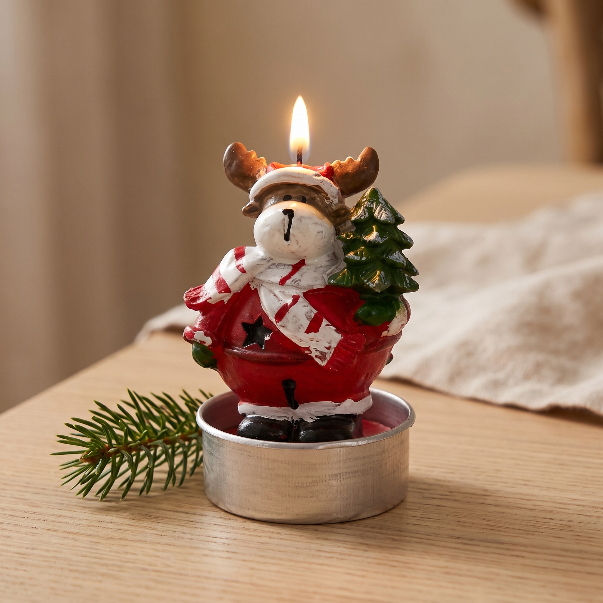 Brennendes Teelicht in Form eines Rentiers mit rotem Weihnachtsoutfit, weißem Schal und Weihnachtsbaum, in Teelicht-Aluhülle, auf Holzoberfläche mit Tannenzweig