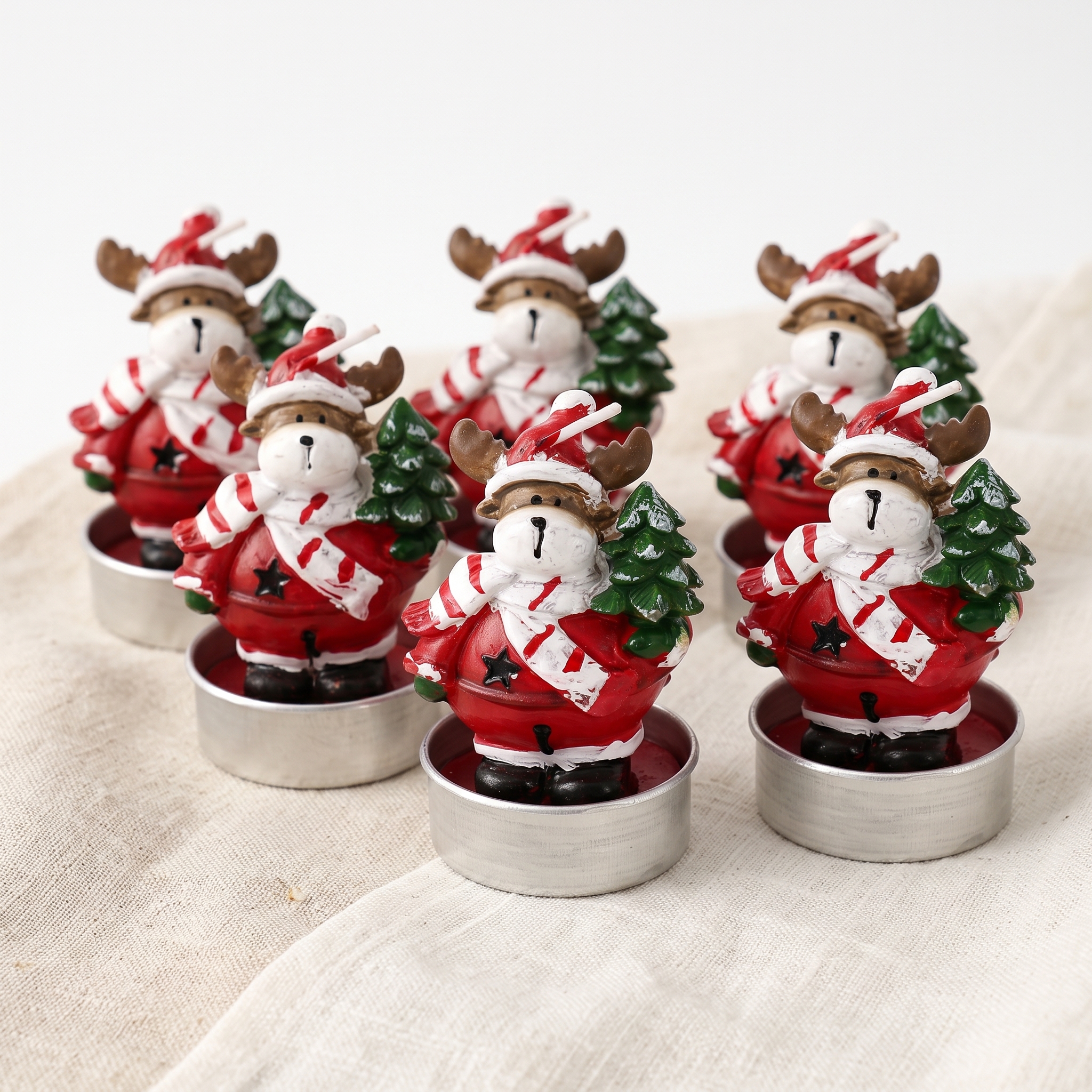 Sechs Teelichter mit weihnachtlichen Elch-Figuren aus Wachs in silbernen Teelichthaltern, im roten Mantel mit rot-weiß gestreiftem Schal und Weihnachtsmütze, einen grünen Tannenbaum haltend, auf cremefarbenem Leinenstoff arrangiert
