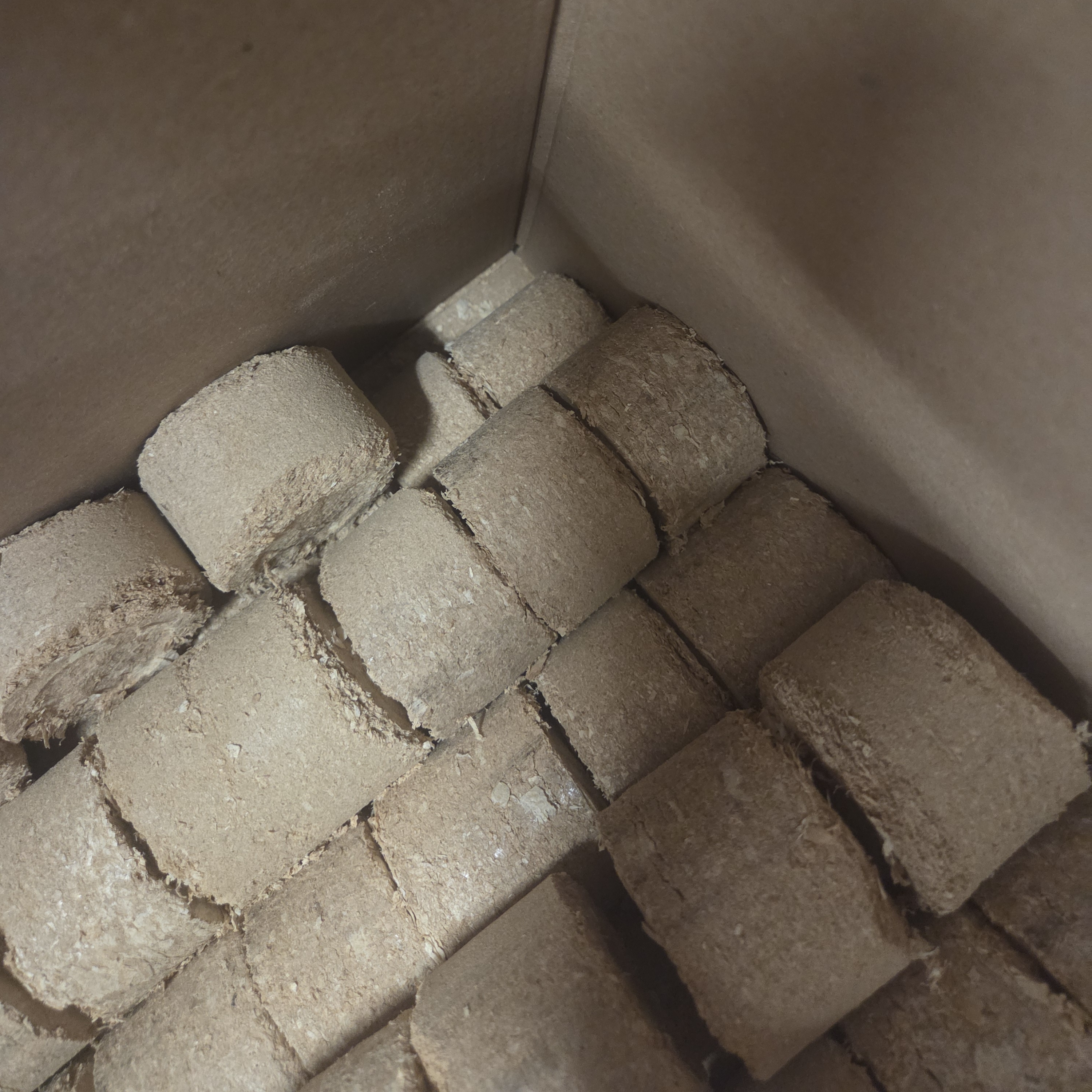 Mehrere beige-braune Holzbriketts aus Schreinerei-Resten gestapelt in einem grauen Karton, Draufsicht in die Ecke des 30-kg-Gebindes