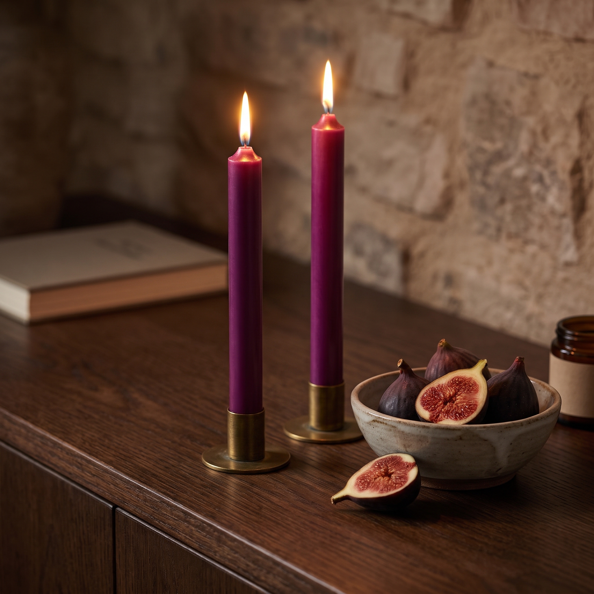 Zwei brennende aubergine-farbene Stabkerzen (180 x 22 mm) in goldenen Kerzenhaltern auf einem dunklen Holzkommode, daneben eine Steinzeugschale mit frischen Feigen vor einer Natursteinwand.