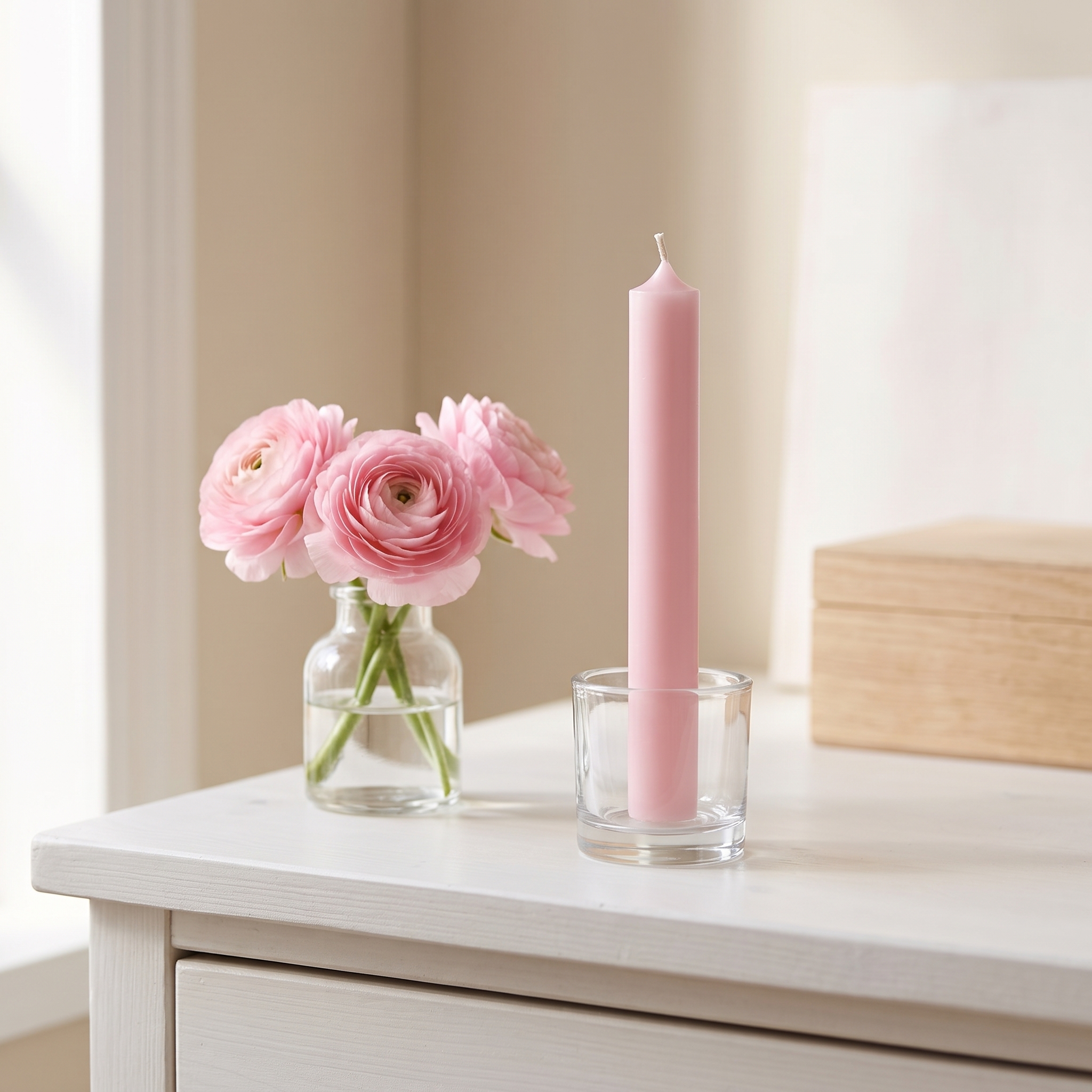Pastellrosafarbene Stabkerze 180 x 22 mm in einem klaren Glas-Kerzenhalter auf weißer Kommode, daneben eine Glasvase mit rosa Ranunkeln
