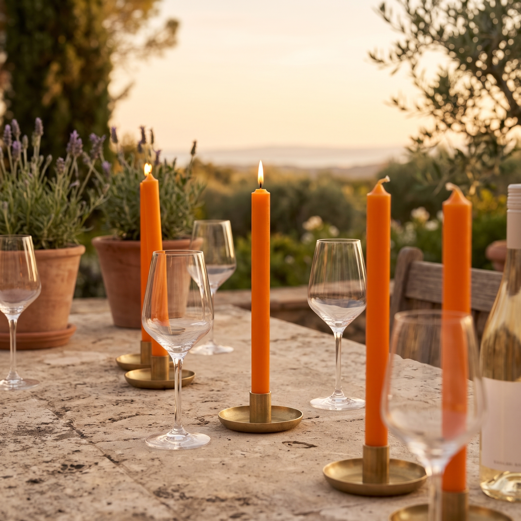 Orangefarbene Stabkerzen 22mm in goldenen Kerzenhaltern auf einem steinernen Tisch im Freien, umgeben von Weingläsern und Terrakotta-Töpfen mit Lavendel im Abenddlicht.