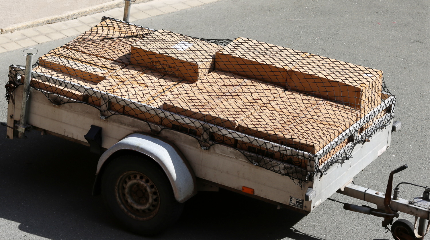 Weißer Einachsanhänger auf Straße, beladen mit Holzplatten, gesichert durch ein beiges Anhängernetz mit schwarzem Randgummi