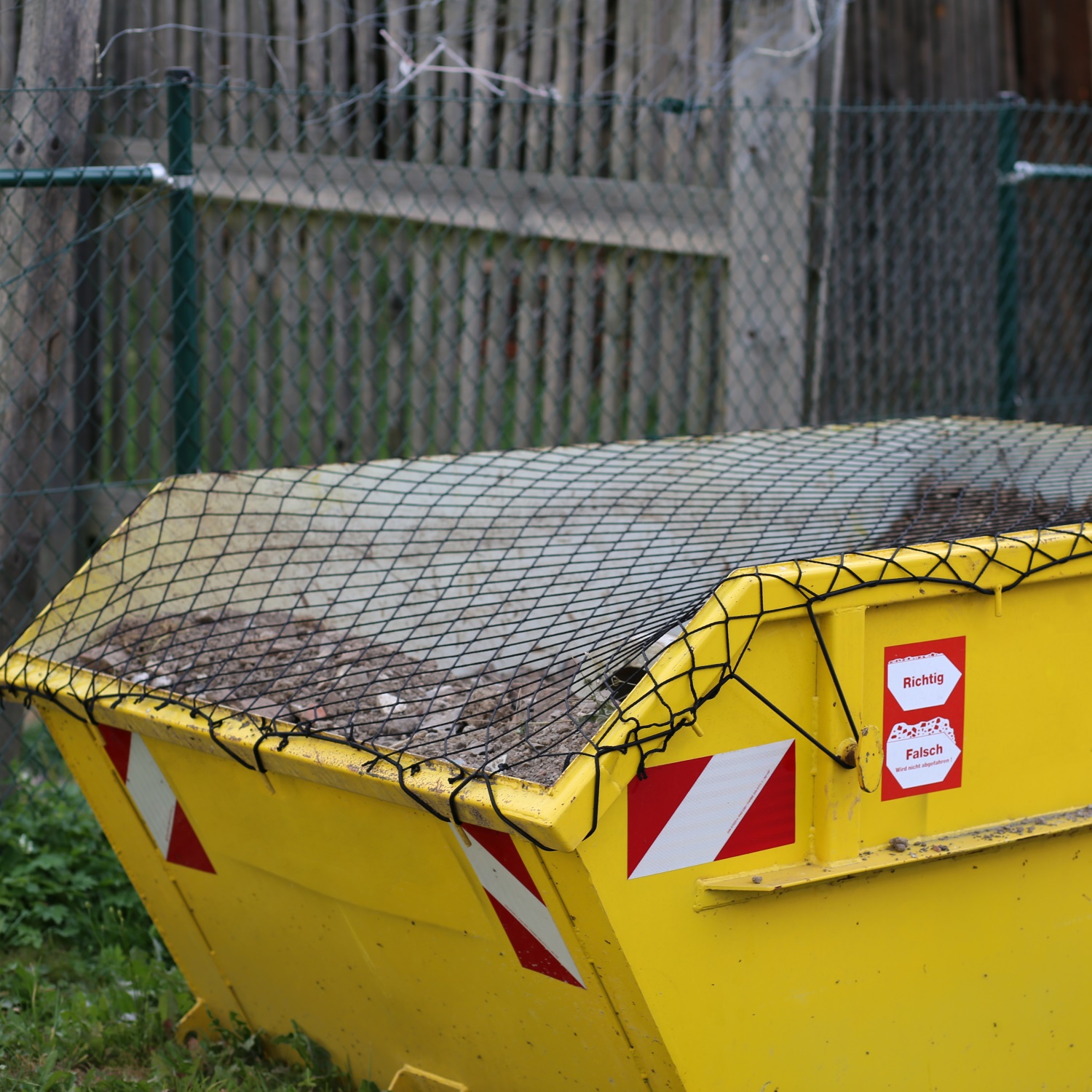 Gelber Baucontainer mit schwarzem Netz abgedeckt, das den Inhalt aus Bauschutt sichert – roter Aufkleber mit Aufschrift „Richtig / Falsch" an der Containerseite, im Hintergrund Maschendrahtzaun und Holzwand