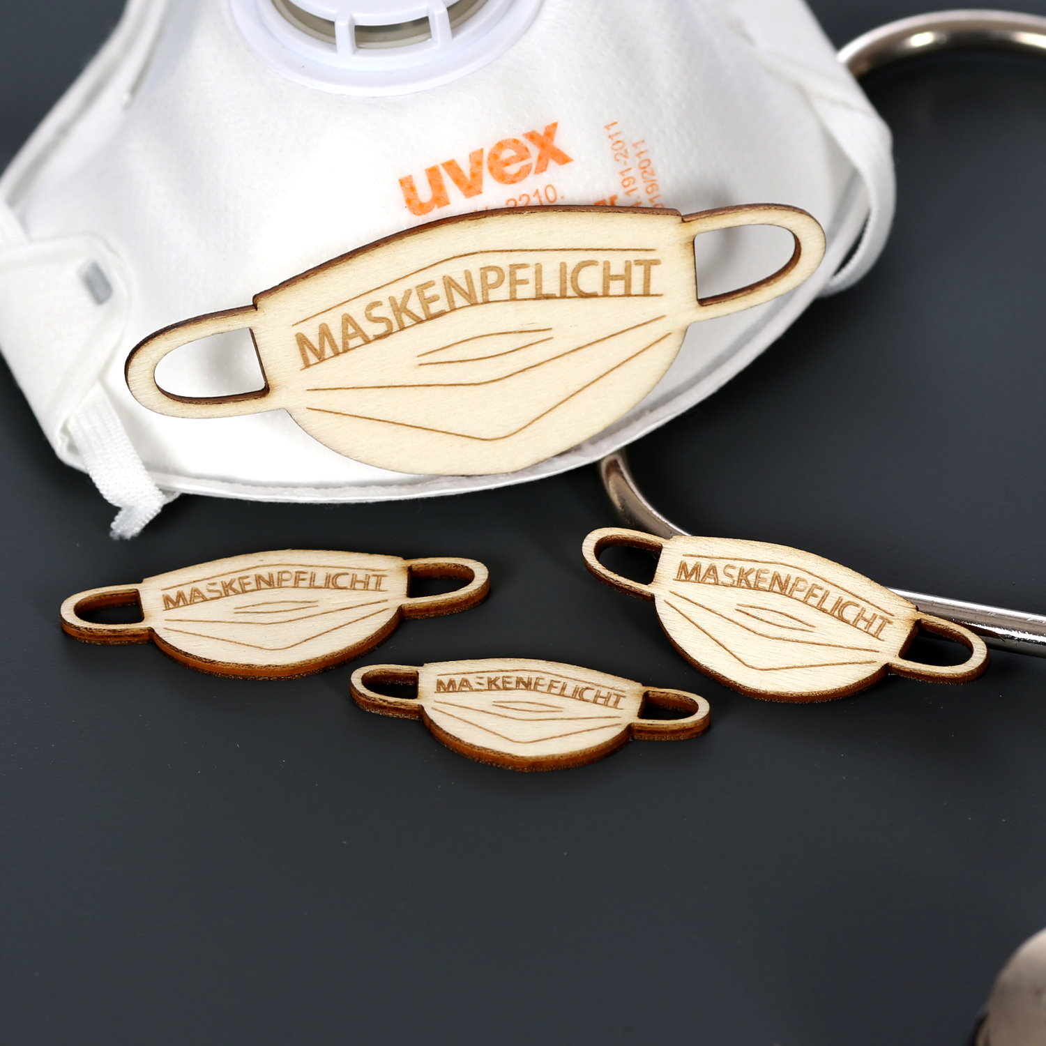Vier Holz-Streuteile in Mundschutz-Form mit Gravur „Maskenpflicht", arrangiert neben einer weißen Uvex-Atemschutzmaske und einem Stethoskop auf grauem Untergrund.